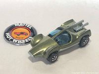 Top side view of metallic green Hot Wheels Mantis A diecast car with blue cockpit and rear engine detailing alongside a round promotional button.