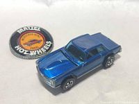 Front-left angle view of blue vintage Mattel Hot Wheels Mercedes-Benz 280SL car with redline wheels and the accompanying circular Mattel Hot Wheels button/badge