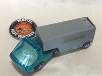 Front angle of the vintage Hot Wheels moving van toy with the blue-green cab, transparent windshield, gray trailer, and collectible button placed beside it showing logo and text.