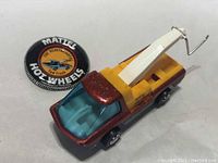 Front-left angled view of the redline tow truck with visible blue windshield and yellow tow crane, alongside the Mattel Hot Wheels button.
