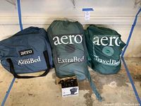 Photo showing three Aero brand air mattresses in their respective bags: Raised AeroBed, ExtraBed, and TravelBed, all twin size. A cardboard box with a battery power pump is visible.
