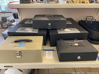 Overall view of all safes stacked on a white table, five SentrySafe safes in black, two smaller safes one beige and one black, all with keys except one beige safe with latch lock.