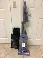 Front view of purple Shark Navigator upright vacuum with attached hose and nozzle beside black storage bag labeled for pet hair clean-up tools.