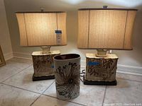 Pair of beige ceramic bird motif lamps with rectangular bases and matching beige lampshades, with a vintage Royal Copenhagen porcelain vase with floral design placed between them on the ground.