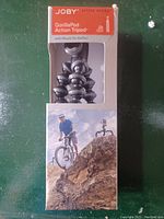 Front view of Joby GorillaPod Action Tripod in original box, showing flexible black legs and mount for GoPro action cameras. Shows product lifestyle photo of tripod mounted on rocky terrain.