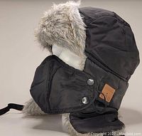 Side profile image of black Russian-style fur-lined winter hat with ear flaps and chin strap, showcasing fuzzy gray faux fur lining and fabric exterior.