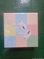 Photo of the front of a sealed retail box displaying white wireless earbuds against a pastel multi-color background.
