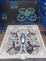 Photo showing Q9s drone in foam packaging with remote control, placed in front of box with product branding and information.