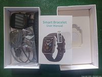 Photo showing a smart watch and smart bracelet in a box with a user manual and charging cables.