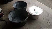 Photo of two galvanized metal basins and one aluminum cooking pot with lid on concrete floor showing overall condition and size.