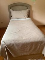 Front view of twin bed with beige bedding covering mattress and upholstered headboard with nailhead trim.