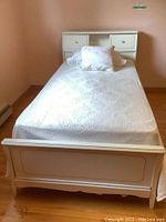 Full view of twin size bed showing white/cream finish with pillows and textured bedspread, featuring a wood headboard with integrated storage drawers and footboard with panel detail.