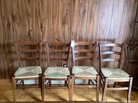 Front view of 4 wooden ladder back chairs with woven reed seats placed against wood panel wall.