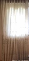 Full photo of one light beige curtain panel with floral embroidery near the bottom edge, sheer fabric allowing light through.