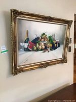 Front view of the still life painting in ornate carved gold frame on wall, showcasing fruit, wine bottle and glasses.