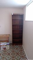 Front view of tall wood veneer shelving unit against light colored wall with six shelves and visible wear.