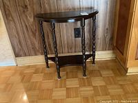 Front angle view of the dark brown wood demi lune table showing turned legs and lower shelf against wood panel wall.