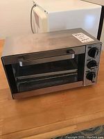 Front and side view of Hamilton Beach toaster oven showing stainless steel body, handle, glass door, and control dials. Tray and metal rack visible inside.