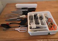Photo showing the cutlery tray with stainless cutlery (forks, spoons, teaspoons), kitchen scissors, and surrounding utensils including knives, whisk, spatula, tongs, and garlic press.