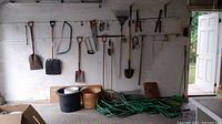 Photo showing multiple garden tools hanging on wall including shovels, rakes, hand saws, hoses, and baskets, with some buckets on floor.