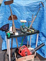 Photo showing assortment of yard tools against blue tarp background including hedge trimmer, leaf blower, rakes, broom, hose nozzle, faucet cover.