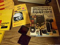 Kodak acetone safelight filter packaging, Kodak projection print scale packet, 'The Handbook of Photographic Equipment' book, and some loose filters displayed on wooden table.