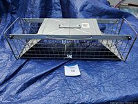 Full view of a rectangular metal humane animal cage with wire mesh sides and two doors, placed on blue tarp for inspection.