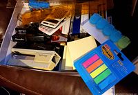Photo of a drawer containing a variety of small office supplies including staplers, calculators, sticky note flags, post-its, rulers, and other small accessories.
