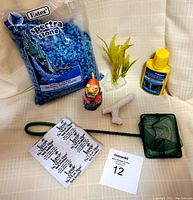 Assorted fish tank supplies photo shows bag of blue Estes Spectra Stone gravel, yellow bottle of aquarium conditioner, artificial yellow plant, decorative orange fish figurine, white coral-shaped stone, green fish net, and pet pet sticker sheet.