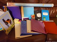 Wide view of laminated world map in tube with assorted folders, binders, composition books, pens, and calculator on brown leather couch.