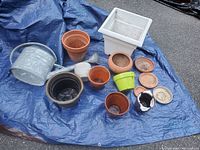 Full lot view showing the metal watering can, white square ceramic pot, assorted terracotta pots, and several ceramic and plastic pots arranged on a blue tarp.
