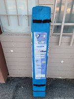 Blue fabric carrying bag for 10ft x 10ft canopy, standing upright against a brick wall, clear label visible with product details and image.