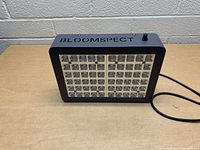 Front angled view of Bloomspect LED grow lamp showing LED bulbs in two sections, power cord, and on/off switch.