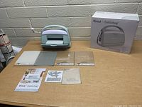 Full lot overview showing Cricut Cuttlebug machine, four plates, instruction manuals, die template, and sample sheet on a table with box in background.