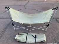 Fold-up hammock set up with visible light green fabric and gray metal frame, carry bag placed on ground nearby.