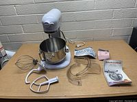 KitchenAid mixer with stainless steel bowl, attachments (whisk, dough hook, flat beater), splash guard, manuals, and power cord on table.
