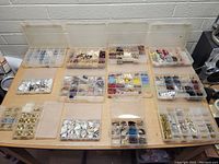 Photo showing a large assortment of plastic compartment organizers filled with beads, shells, hooks, and other jewelry making components arranged on a table.