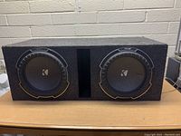 Front view of dual Kicker 12 inch subwoofers in carpeted enclosure showing both cones and center port.