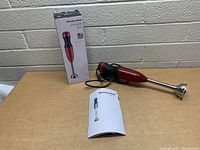 KitchenAid hand blender with cord, original box, and user manual on table.