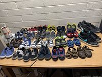 Photo showing various kids shoes in assorted colors and styles arranged in rows on a table with a brick wall background.