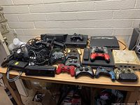 Full overview of consoles, controllers, accessories on wooden table against wall