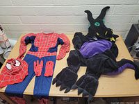 Photo showing the Spiderman costume laid flat with gloves and mask, and the dragon costume with headpiece, tail, and clawed gloves.