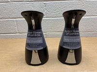 Two black Better Homes & Gardens automatic soap dispensers side by side with labels visible, placed on a table against a white brick background.