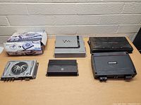 Five car stereo amplifiers arranged on a wooden table, visible are a silver Vision amplifier, black Kicker amplifier, black JBL amplifier GX-A660, smaller black amplifier, and a silver amplifier with spider logo in original box.