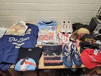 Five folded tops (four graphic T-shirts and one embroidered jacket), three caps and two pairs of shoes on a table
