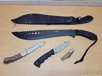 Top-down view of the large black serrated machete alongside three different folding knives on a wooden surface.