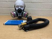 Front angle photo of the MSA Comfo Classic respirator mask fitted with dual pink filter cartridges in metal housing, flexible black hose with brass connector and a box labeled MSA Cover Lens for Ultravue faceniece.