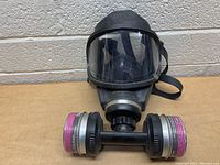Front view of black MSA gas mask showing clear face shield and dual filters attached by central tube.