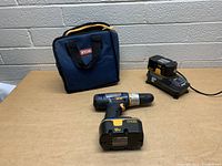 Overall view of Ryobi 18V cordless drill, two batteries, charger, and carrying bag on a brown surface.