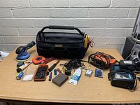 Front view of tool bag with assorted tools arranged on table including orbital sander, jigsaw, measuring tape, bit set, battery chargers, wire cutters, tubes, and various hand tools and electrical accessories.